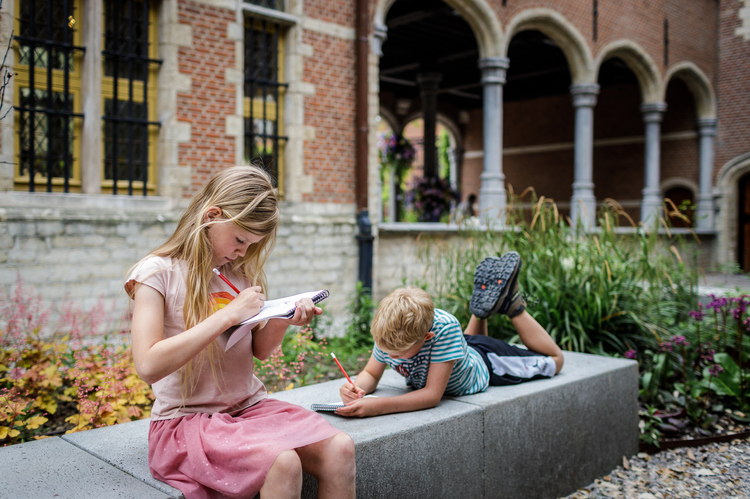 Twee kinderen zitten op een bank in de museumtuin. Ze zijn in een boekje aan het schrijven of tekenen.