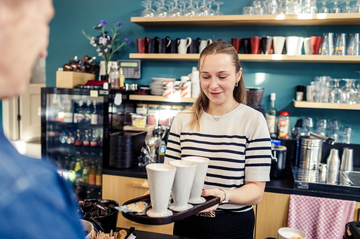 Iemand in een gestreepte trui overhandigt al glimlachend een plateau met kopjes koffie.