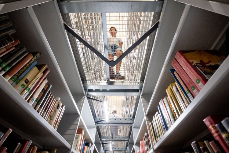 De foto is genomen vanuit een laag perspectief en naar boven gericht. Door een metalen rastervloer zie je een persoon staan. Aan beide kanten staan rekken gevuld met boeken.