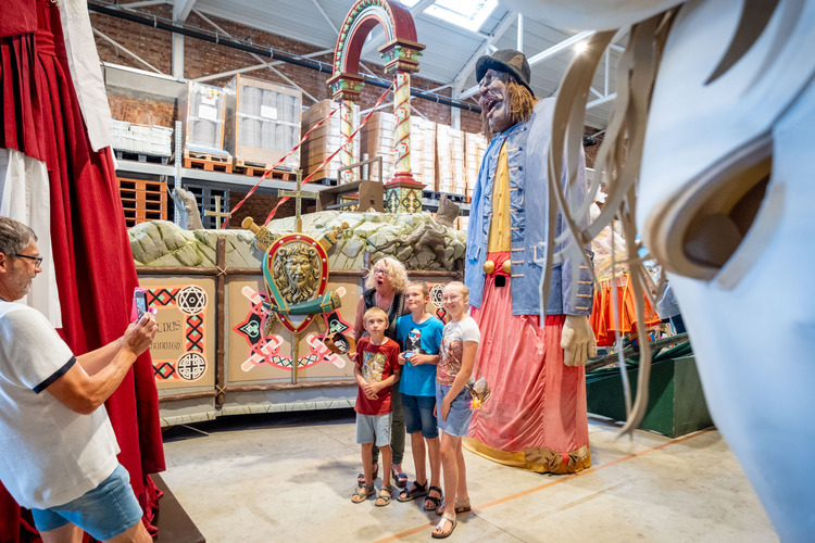 Een vrouw poseert met drie kinderen voor één van de reuzen in een loods. Je ziet impressionante praalwagens rondom hen.