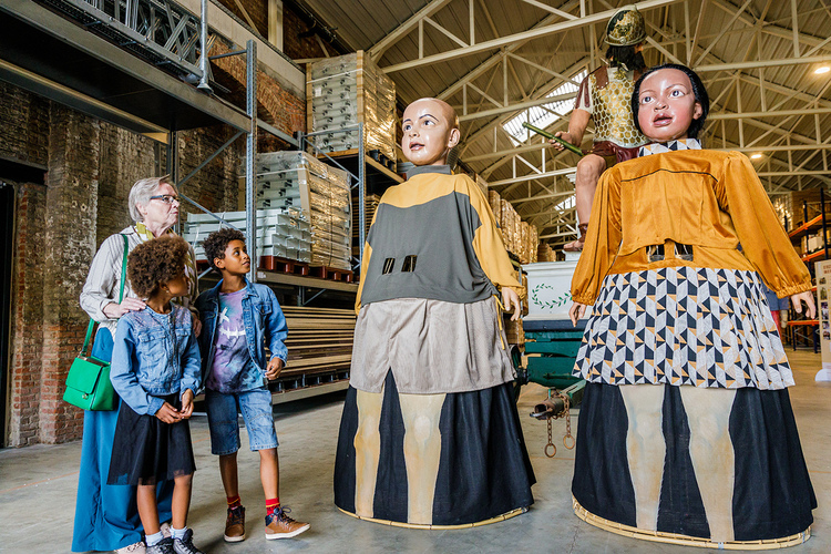 Een volwassene en twee kinderen staan in een loods waar twee van de Mechelse reuzen staan.