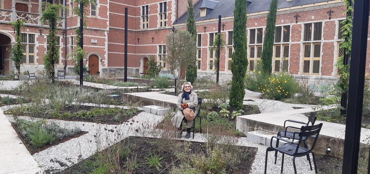 Een glimlachende dame zit op een tuinstoel in de museumtuin. De paden tussen de plantenbakken vormen een ruit-patroon.